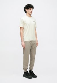 Göndör hajú férfi állva, bézs rövid ujjú logós pólót, taupe színű melegítőnadrágot, fekete edzőcipőt és fekete zoknit visel fehér háttér előtt.