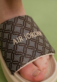 Fod i en beige og brun Air Jordan slide sandal med et mønstret rem og lysegrøn baggrund.