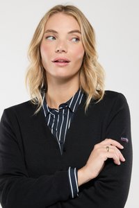 Cardigan nero con bottoni, lavorazione a maglia testurizzata, indossato sopra una camicia a righe blu navy. Accentatura del logo ben visibile sulla manica. Modella in posa con le braccia incrociate.