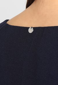 Dunkelblaues Kleidungsstück mit glatter Textur; verfügt über ein rundes silbernes Etikett am hinteren Halsausschnitt. Einfaches, minimalistisches Design.