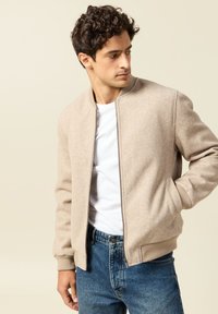 Beige bomber jacket in a soft texture, featuring a zip front, ribbed cuffs, and side pockets, worn over a white t-shirt and blue jeans.