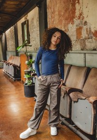 Blauw gestreepte lange mouwen top, beige baggy broek en witte chunky sneakers. Staande in een industriële ruimte met houten vloeren en leren stoelen.