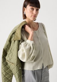 Lichtgroene gewatteerde jas gecombineerd met een crèmekleurige textuurblouse met een ronde hals en knopen. Model toont oversized, opgeblazen mouwen.