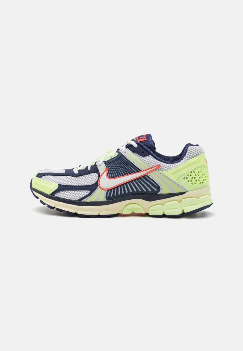 Ténis desportivos com parte superior em malha nas cores azul marinho, branco e amarelo neon, com um swoosh vermelho em contraste, sola almofadada e detalhes texturizados.