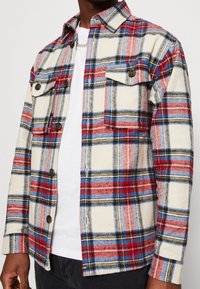 Czerwono-niebieska i beżowa koszula w kratę z guzikami, z dwiema kieszeniami na piersi. Wykonana z miękkiego materiału, z luźnym fasonem i zapięciem na guziki.