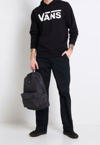 Felpa nera con logo "VANS" bianco, pantaloni scuri e scarpe slip-on nere. Tiene uno zaino nero a trama con una tasca frontale.