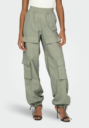 Pantalon cargo ample vert clair avec taille élastique et liens aux chevilles, porté avec des sandales noires à talons hauts et un top blanc.