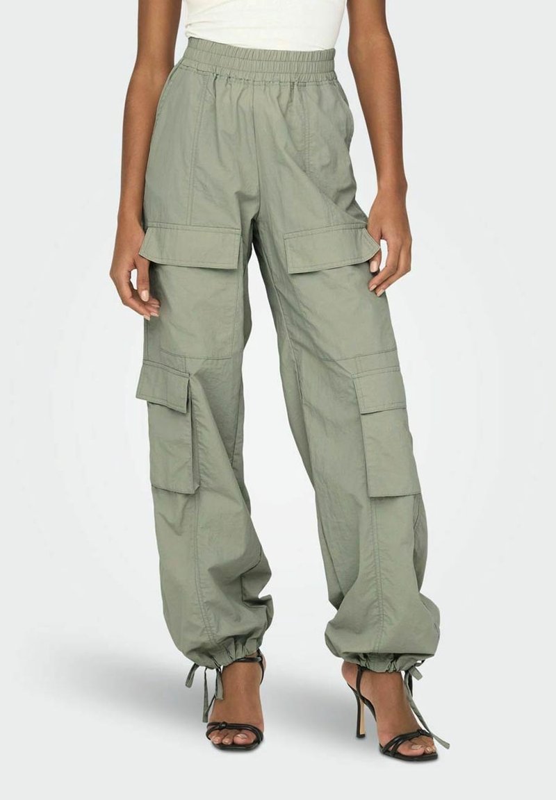 Pantalon cargo ample vert clair avec taille élastique et liens aux chevilles, porté avec des sandales noires à talons hauts et un top blanc.