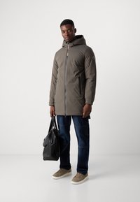 Padded olive-green parka with a zip front, hood, two side pockets, worn with blue jeans and beige sneakers. Black bag held in the hand.