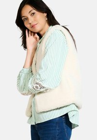 Creame vest van fleece met een groene en witte gestreepte blouse met kantaccenten op de mouwen, gedragen met blauwe jeans.