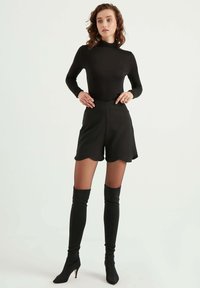 Zwarte bodysuit met lange mouwen, gecombineerd met hoge zwarte shorts met een geschulpte zoom. Model draagt kniehoog zwarte hoge hakken.