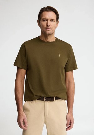 Uomo con t-shirt verde oliva a maniche corte e pantaloni beige, in piedi contro uno sfondo chiaro uniforme, rivolto verso avanti.