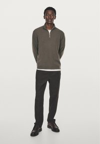 Zip-neck tröja i olivgrön, tillverkad av mjukt material. Parat med mörka byxor och bruna skor, stående mot en enkel bakgrund.