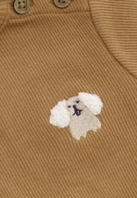 Beige geripptem Stoff mit einem kleinen gestickten weißen Hund mit flauschigen Ohren und marineblauen Gesichtszügen neben zwei beigen Knöpfen.