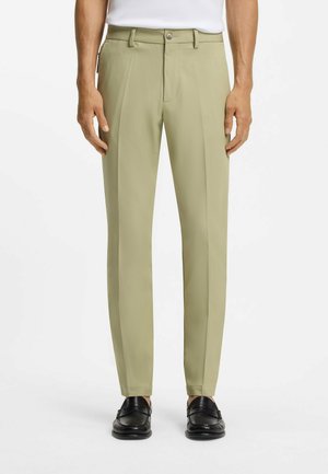 Hombre con pantalones beige a medida y mocasines negros, de pie frente a un fondo blanco, con las manos relajadas a los lados.