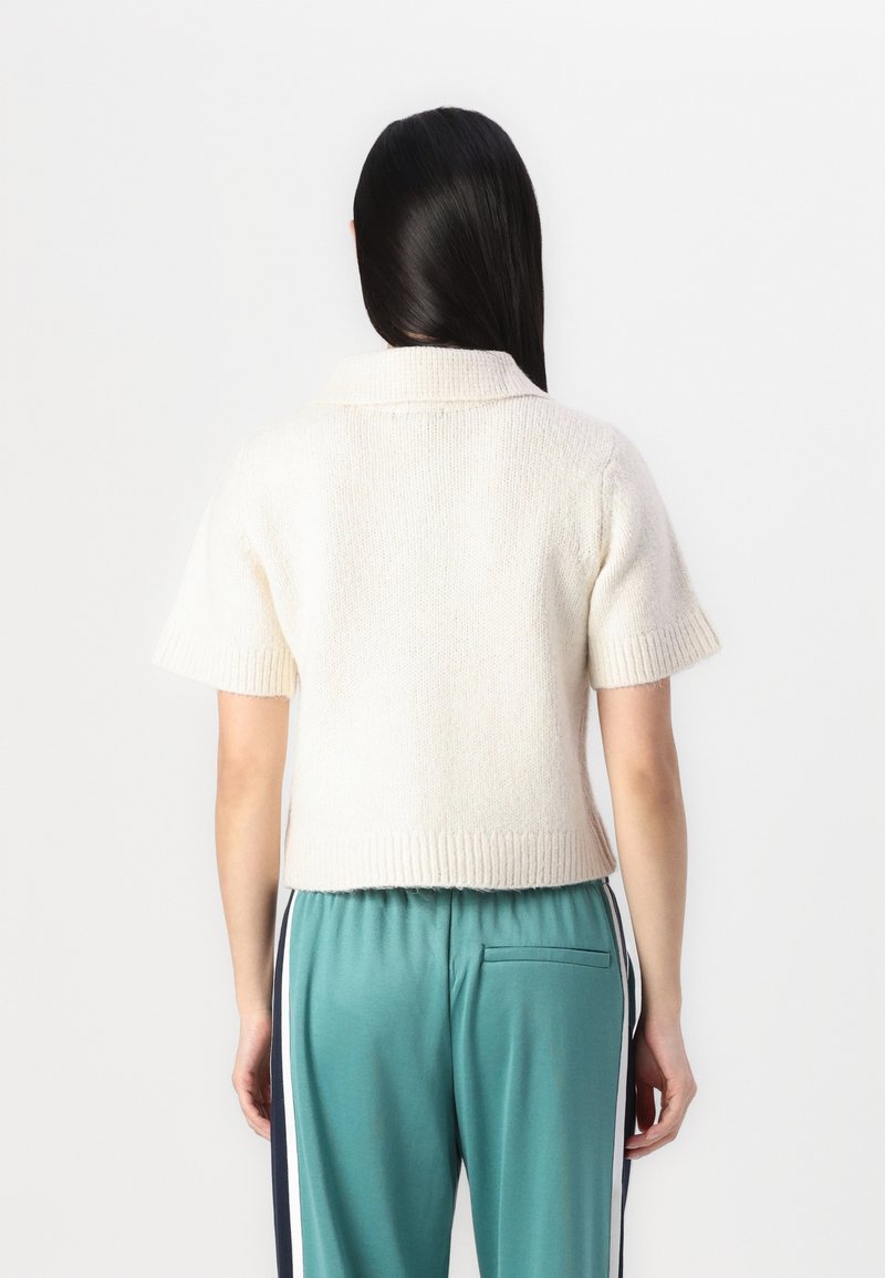 Cremfarbener Kurzarmpullover mit geripptem Saum und Kragen, strukturiertem Stoff. Kombiniert mit türkisfarbenen Hosen mit Seitenstreifen und einer Gesäßtasche.