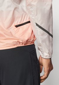Lett, semi-transparent jakke i gradient rosa-til-hvit stoff, med en glidelåslomme og svarte detaljer; elastiske mansjettdetaljer synlige.