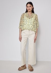 Helles gelbes Kurzarm-Bluse mit floralem Muster und Schnürkragen, kombiniert mit cremefarbenen, weit geschnittenen Hosen und hellbraunen Slipper-Sandalen.