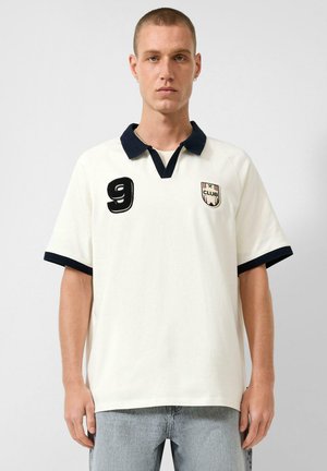 Junger Mann mit kurzen Haaren, der ein weißes Poloshirt mit schwarzem Kragen und Ärmelsäumen, Nummer 9 auf der Brust und hellblaue Jeans trägt.