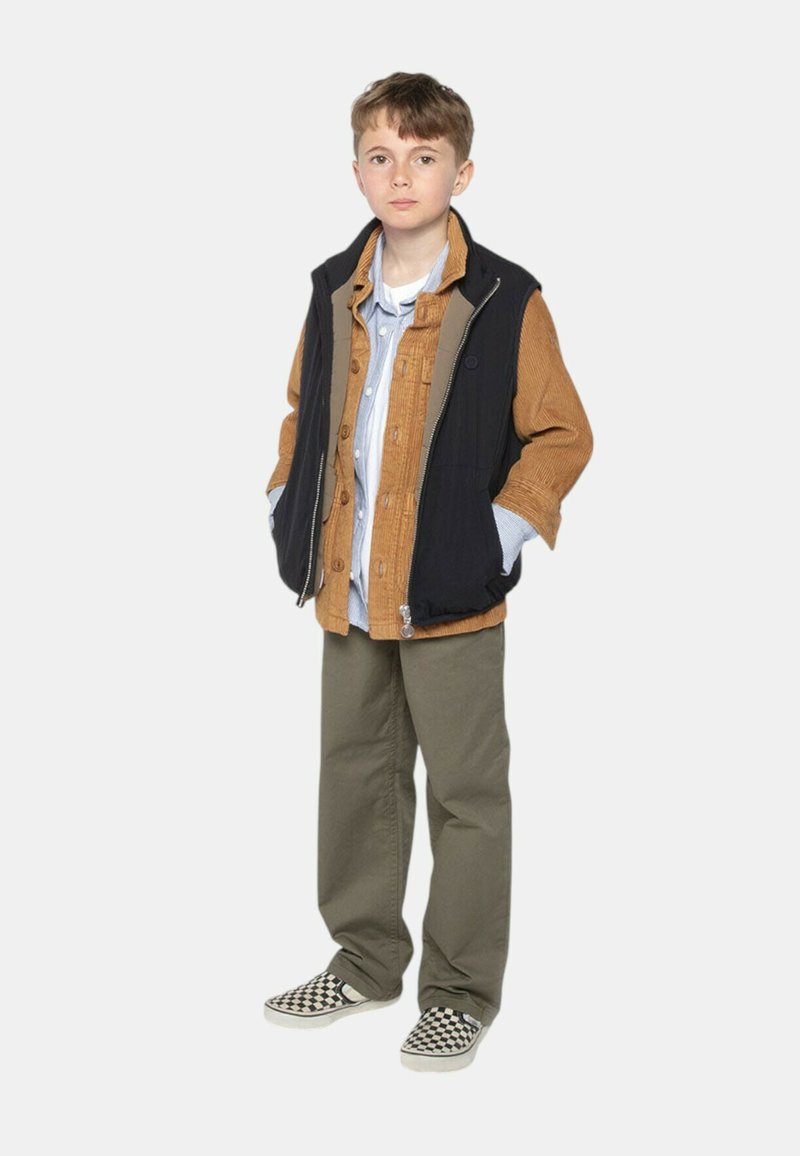 Tenue jeune avec un gilet noir, une chemise en velours côtelé marron, une chemise bleue à boutons, un pantalon vert olive et des chaussures à damier noir et blanc.