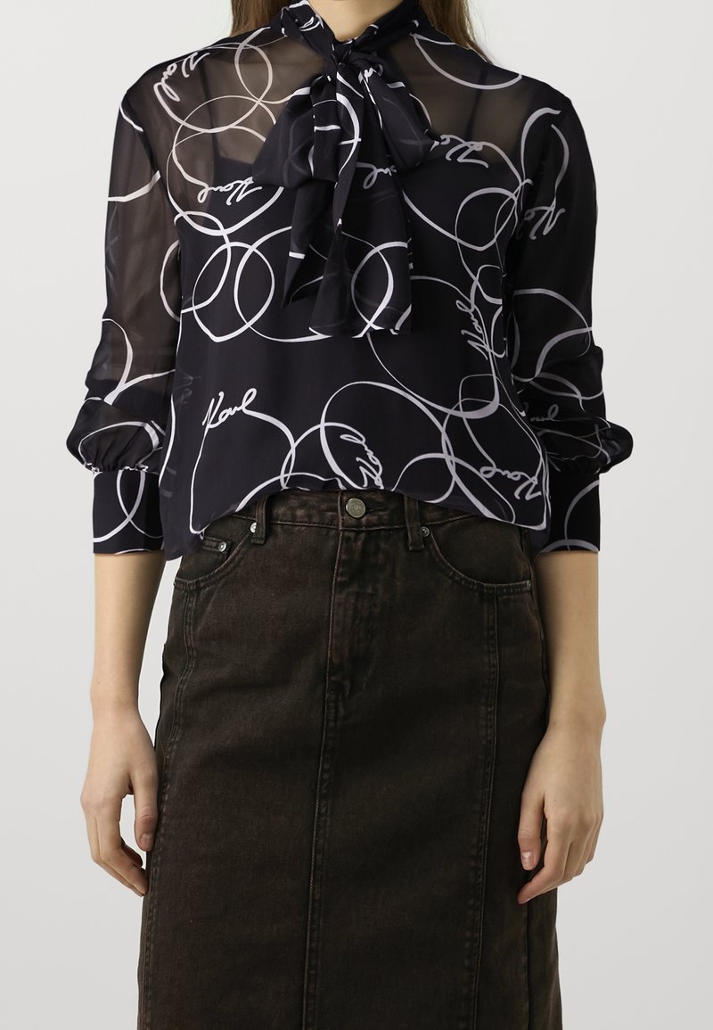 Femme portant un chemisier noir transparent avec des motifs de lignes abstraites blanches et un col à nœud papillon, associé à une jupe en jean foncée taille haute.