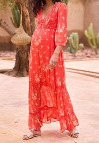 Femme enceinte portant une longue robe rouge à fleurs et des sandales beiges, debout à l'extérieur près d'arbres et de cactus.