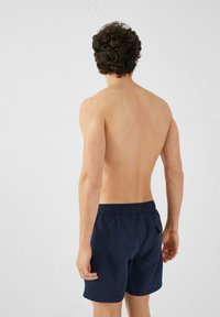 Pantaloni da bagno blu navy con elastico in vita e una tasca posteriore, visti dalla schiena di una figura maschile. Texture del tessuto liscia.