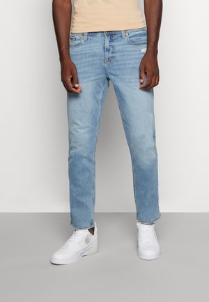 Mann trägt hellblaue Jeans und weiße Turnschuhe, steht vor einem einfarbigen, hellen Hintergrund.