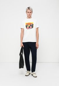 White cotton t-shirt with colorful graphic print, navy trousers, and a black shoulder bag; model wears cream sneakers with dark stripes.