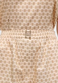 Tissu beige avec un motif répétitif de rectangles et cercles aux contours marron, présentant une taille élastique ornée d'un petit logo crocodile cousu.