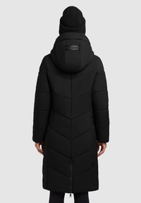 Abrigo acolchado negro con costura en forma de chevrón, capucha y logotipo en la parte posterior. Cuenta con un cierre frontal y mangas largas. Se lleva con un gorro de punto negro.