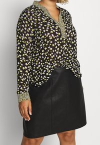 Blouse noire à motif floral jaune et blanc, patte de boutonnage, et col à motif contrastant. Associée à une jupe en cuir noire élégante.