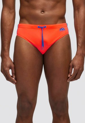 Torace e parte superiore delle gambe maschili che indossano slip da bagno arancioni con coulisse blu e logo sul lato destro contro uno sfondo chiaro e uniforme.