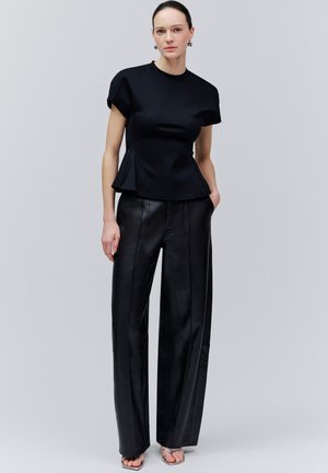 Femeie cu păr închis la culoare, purtând un top negru cu mâneci scurte și pantaloni negri largi, stând cu o mână în buzunar, pe un fundal simplu gri.