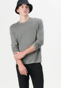 Grå långärmad skjorta i ett mjukt tyg med rund halsringning och upprullade ärmar, kombinerad med svarta byxor och en svart bucket hat.