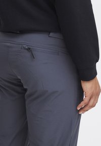 Mørk grå, letvægtsbukser med en lynlås baglomme og glat tekstur. Livremmen er elastisk, designet til komfort og fleksibilitet.