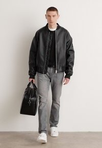 Chaqueta bomber negra de piel sintética con cierre de cremallera, llevada sobre una camiseta negra, combinada con jeans grises y zapatillas blancas, sujetando un bolso negro.