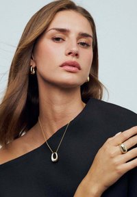 Guldsmykkesæt med en pendantsmykke med oval design, hoopsølvøreringe og en tekstureret ring, båret mod en sort stofbaggrund.