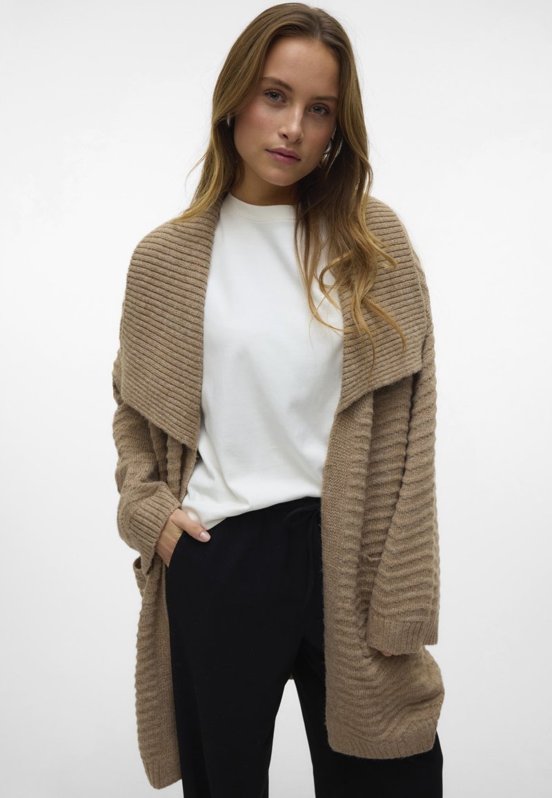 Beige geripptes Cardigan mit offener Vorderseite, langen Ärmeln und strukturiertem Strick. Kombiniert mit einem weißen Oberteil und schwarzen Hosen.