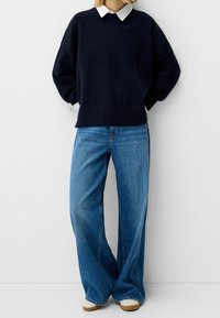 Pull en tricot bleu marine avec un ourlet côtelé porté par-dessus une chemise blanche à col, associé à un jean denim bleu à jambes larges et des chaussures de couleur claire.