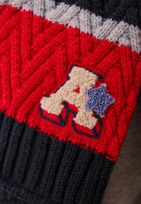 Knit garment featuring red and navy yarn with a textured zigzag design. An embroidered beige letter "A" with a blue star accent is prominent.