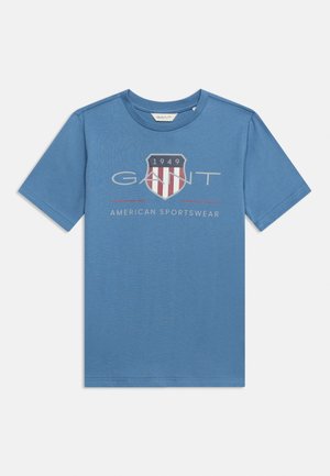Μπλε κοντομάνικη μπλούζα με λογότυπο ασπίδας που αναγράφει "1949", τη λέξη "GANT" και "American Sportswear" τυπωμένα στο στήθος.