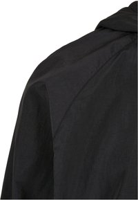 Veste noire en tissu lisse et texturé, avec une capuche et des détails de couture visibles sur les épaules et le haut du dos.