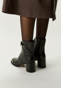 Kobieta nosząca czarne skórzane botki za kostkę na słupku, czarne przezroczyste rajstopy i brąskową skórzaną spódnicę na białym tle.