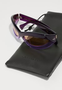 Ochelari de soare violeți cu o formă unică de ochi de pisică, având lentile gradient și un toc neted de culoare neagră. Accentele includ branding pe brate.