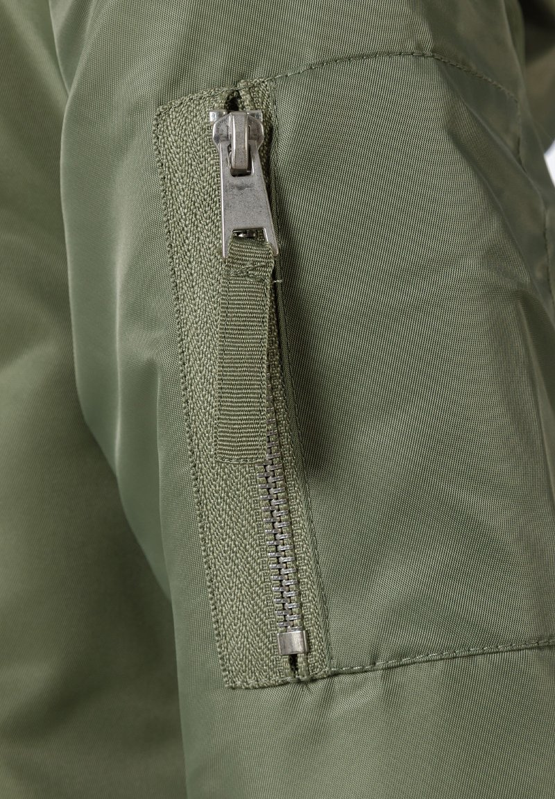 Primer plano de una chaqueta verde olivo con cierre de cremallera metálico y tirador de tela, que presenta una superficie texturizada y costuras reforzadas.