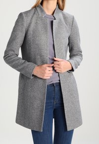 Abrigo de lana gris con mezcla de tejidos, con cuello alto, corte ajustado y diseño de abertura frontal. Combinado con una camisa morada de canalé y jeans de mezclilla.