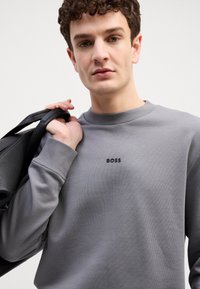 Hallitav hall sweatshirt, millel on ribbed kaksteist ja must "BOSS" logo rinnal. Mudel hoiab käes tekstuuriga musta kotti, näidates sujuvat kangastekstuuri.