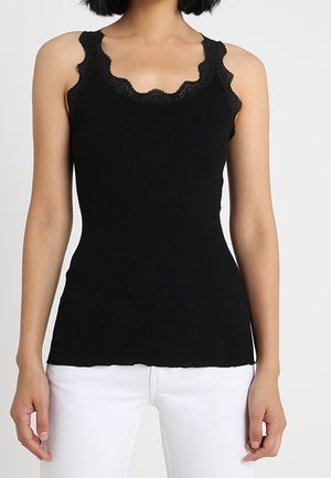 Nainen pukeutuneena mustaan ribbineulottuun toppiin, jossa on pitsireunus pääntiellä, ja valkoiset housut, seisoo yksivärisen taustan edessä.