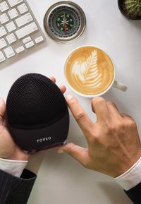 Brosse de nettoyage facial en silicone noir avec une surface texturée, tenue dans une main à côté d'un latte avec un motif de feuille, d'une boussole et d'un clavier.